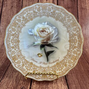 May include: A decorative porcelain plate featuring a central illustration of a white rose with green leaves. The plate has a scalloped edge with a gold filigree border and the text "Diana, A Rose Everlasting" printed in gold script at the bottom.