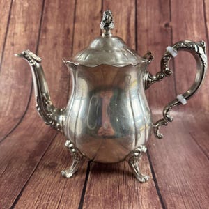 May include: Silver-colored teapot with ornate detailing. The teapot features a curved spout, a decorative handle, and a lid with a sculpted finial. The teapot is set on three small feet. The background is a wooden surface.
