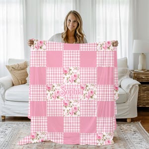 Może przedstawiać: Różowa narzuta patchwork z kwiatowym wzorem róż i imieniem "Sophia" w ozdobnej czcionce. Narzuta ma wzór w kratę vichy i jednolite różowe kwadraty. Narzuta jest trzymana przez osobę.