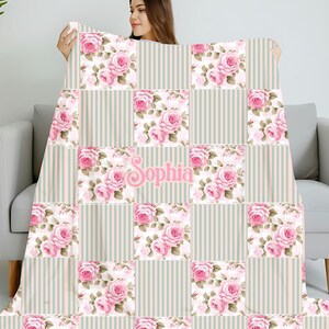 Puede incluir: Una manta personalizada con un diseño de patchwork. Presenta cuadrados alternos de rosas rosas y rayas verticales. El nombre "Sophia" se muestra en escritura rosa. La manta se sostiene, mostrando su suave patrón floral.