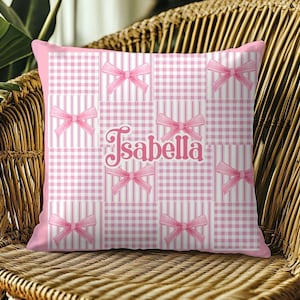 May include: Pink decorative pillow with a patchwork design. The pillow features a mix of gingham and striped patterns, adorned with pink bows. The name "Isabella" is scripted in pink across the center.