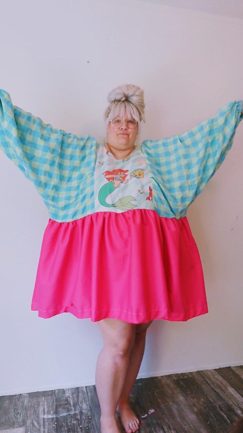 May include: A bright pink and blue dress with a cartoon mermaid graphic. The top features a blue and green checkered pattern, while the skirt is a vibrant pink. The sleeves are wide and flowing.