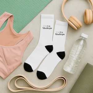 Può includere: Un'immagine piatta con accessori per il fitness. Calzini bianchi con talloni neri e la scritta "Simply Liv Mockups" accanto a un reggiseno sportivo color pesca, una bottiglia d'acqua e cuffie beige. Sono visibili anche un tappetino da yoga verde e una fascia elastica.