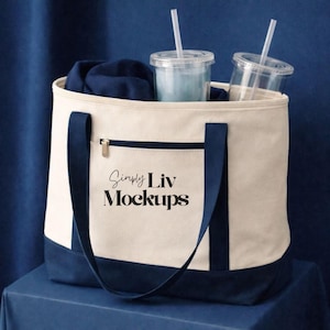 May include: A canvas tote bag with a cream-colored body, navy blue accents, and a zippered pocket. The bag contains two clear plastic cups with straws and a navy blue garment. The text "Simply Liv Mockups" is printed on the front.