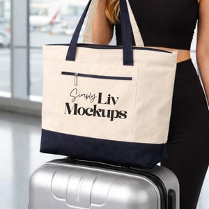 Può includere: Borsa tote color crema con dettagli blu navy, una tasca con cerniera e manici blu navy. Il testo "Simply Liv Mockups" è stampato sul davanti. La borsa è appoggiata su una valigia argentata.