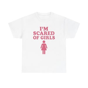 Può includere: T-shirt bianca con la scritta rosa "I'M SCARED OF GIRLS" e un disegno di un personaggio rosa. Il testo e il disegno sono centrati sulla parte anteriore della maglietta. La maglietta è probabilmente in cotone o in un materiale simile.