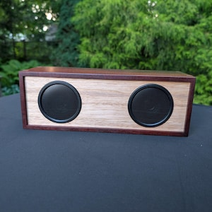 May include: A rectangular wooden speaker featuring a dark brown frame and a light-coloured wood front panel. The device includes two circular black speaker grilles positioned side-by-side on the front face, creating a clean and functional design.