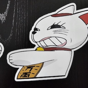 May include: A white cartoon cat sticker with a red collar and accents, a gold bell, and a gold plaque with Japanese text. The cat has an angry expression and is holding up its paw. The sticker is on a black surface.