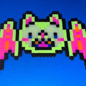 May include: Pixel art bat made from colorful beads. The bat features a light green body, pink wings, and black outlines. The bat's face has blue eyes, a pink nose, and a black mouth. The background is a solid blue.