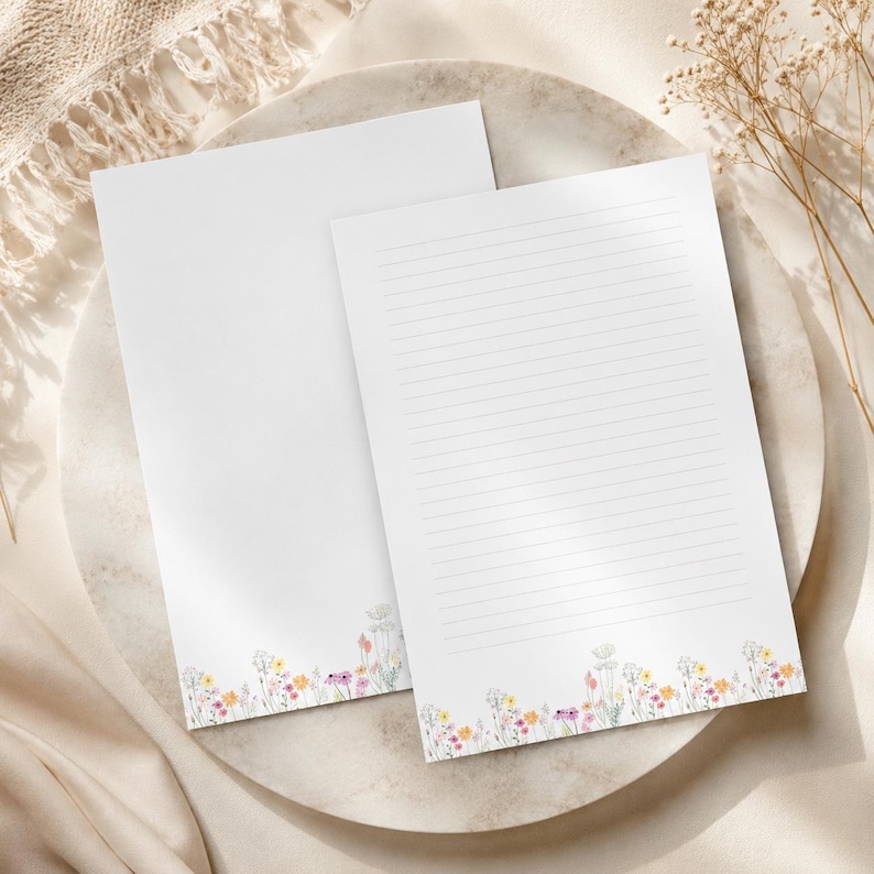 K&ouml;nnte beinhalten: Zwei Blatt wei&szlig;es Briefpapier, eines unbedruckt und eines mit horizontalen Linien. Beide Bl&auml;tter zeigen am unteren Rand eine zarte, farbenfrohe Illustration von Wildblumen in den Farben Gelb, Rosa und Violett auf dem Papier.