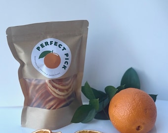 May include: A kraft paper bag with a clear window showing dehydrated orange slices. A whole orange and dried orange slices are arranged near the bag. The bag has a circular label that reads "Perfect Pick" with an orange illustration.