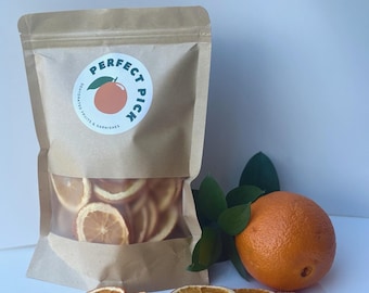 May include: A brown paper bag with a clear window filled with dried orange slices. The bag has a sticker that says "Perfect Pick." Next to the bag are fresh orange slices, a whole orange, and green leaves. The product is dehydrated fruits and garnishes.