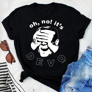 May include: Black t-shirt with the text "oh, no! it's" above a white graphic of a person covering their eyes and mouth. Below the graphic is the word "DEVO" in white block letters. Sunglasses and jeans are visible.