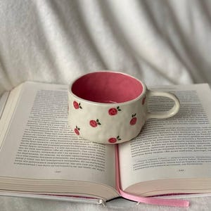 Peut inclure: Une tasse en céramique blanche ornée de roses rouges et d'un intérieur rose, posée sur un livre ouvert avec un ruban rose. Le fond est un tissu blanc et doux. Idéal pour déguster une boisson chaude.