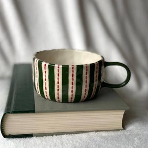 Peut inclure: Une tasse en céramique avec une anse verte et un intérieur blanc. La tasse est ornée de rayures vertes verticales et de lignes rouges pointillées. Elle repose sur un livre fermé avec une couverture verte.