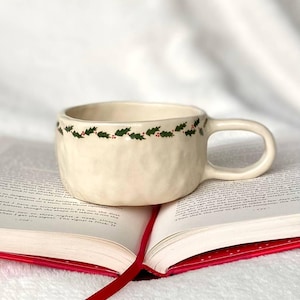 May include: Cream ceramic mug with a textured finish, decorated with a green holly and red berry pattern around the rim. The mug rests on an open book with a red ribbon bookmark, creating a cosy, festive scene.