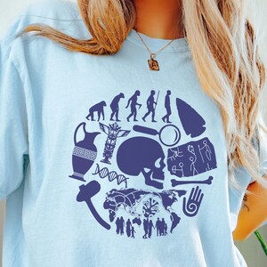 May include: A light blue t-shirt featuring a circular dark blue graphic print. The design includes an evolution sequence, a skull, a Greek-style vase, a totem pole, a magnifying glass, a stone tool, a DNA strand, a map, and cave art figures.