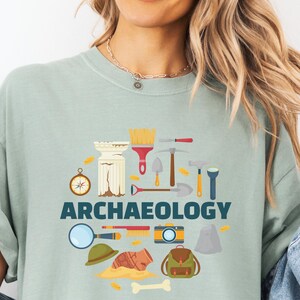 Op de afbeelding: Een saliegroen t-shirt met een grafische print met de tekst ARCHAEOLOGY in donkerblauw. Het ontwerp bevat illustraties van een kompas, een stenen zuil, een borstel, een troffel, een houweel, een hamer, een zaklamp, een vergrootglas, een camera, een hoed, een rugzak, een potscherf en een bot.
