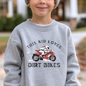 Puede incluir: Sudadera gris claro con un gráfico de una moto de cross roja y blanca y un piloto. El texto "THIS KID LOVES DIRT BIKES" está impreso encima y debajo del gráfico en negro.