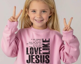 May include: Pale pink sweatshirt featuring the words "LOVE JESUS" and the Bible verse John 13:34. The sweatshirt also has the text "A new command I give you: Love one another. As I have loved you, so you must love one another."