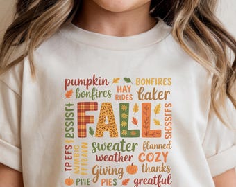 Puede incluir: Una camiseta beige con un diseño colorido con temática otoñal. La palabra "FALL" en el centro, en un patrón de cuadros, rodeada de palabras como "pumpkin", "bonfires" y "cozy". La camiseta la lleva una persona.