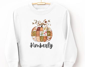 Puede incluir: Sudadera blanca con un diseño de calabaza de retazos en colores otoñales y detalles florales. El nombre "Kimberly" está escrito en cursiva debajo de la calabaza. La sudadera está colgada en una percha de madera.
