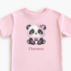 Peut inclure: T-shirt rose clair avec un dessin de panda. Le panda est noir et blanc avec un nœud rose et le nom "Florence" en dessous. Le t-shirt est de couleur rose uni.