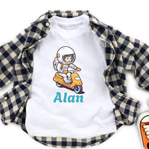 Könnte beinhalten: Weißes T-Shirt mit einem Cartoon-Astronauten auf einem gelben Roller. Der Name "Alan" steht türkis darunter. Ein schwarz-weiß kariertes Hemd ist darüber gelegt. Orangefarbene und weiße Turnschuhe sind zu sehen.