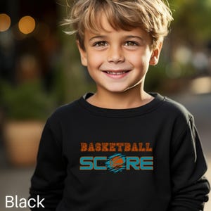May include: A black sweatshirt featuring the words "BASKETBALL SCORE" in orange and teal. The sweatshirt is worn by a young child with blond hair, smiling at the camera. The word "Black" is written at the bottom.