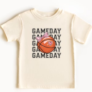 May include: A cream-coloured t-shirt with a basketball graphic, a pink bow, and a heart. The word "GAMEDAY" is printed in a stacked design. The shirt is made of a soft material.