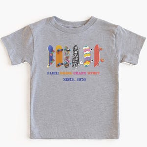 May include: Athletic heather gray t-shirt featuring a row of colorful skateboards. The shirt has the text "I LIKE DOING CRAZY STUFF SINCE 1970" in a rainbow font. A casual tee for kids.