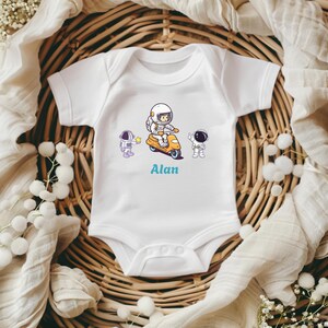 Könnte beinhalten: Weißer Baby-Body mit einem Cartoon-Astronauten-Design. Das Design zeigt einen Astronauten auf einem gelben Roller, mit zwei weiteren Astronauten und dem Namen "Alan" in Blau. Der Body ist aus weichem Material.