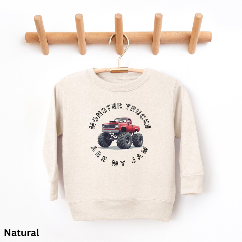 Puede incluir: Una sudadera de color natural con un gr&aacute;fico de un cami&oacute;n monstruo rojo y el texto "MONSTER TRUCKS ARE MY JAM" en un dise&ntilde;o circular. La sudadera est&aacute; colgada de un perchero de madera.