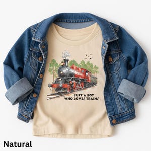 May include: A natural-coloured t-shirt featuring a red and black steam train graphic, the text "JUST A BOY WHO LOVES TRAINS", and a blue denim jacket. The train is on tracks with trees and birds in the background.