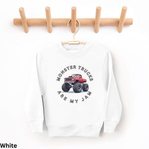 Puede incluir: Sudadera blanca con un gr&aacute;fico de cami&oacute;n monstruo rojo. El gr&aacute;fico presenta un cami&oacute;n rojo con neum&aacute;ticos negros grandes y el texto "Monster Trucks Are My Jam" en un dise&ntilde;o circular. La sudadera est&aacute; colgada en una percha de madera.