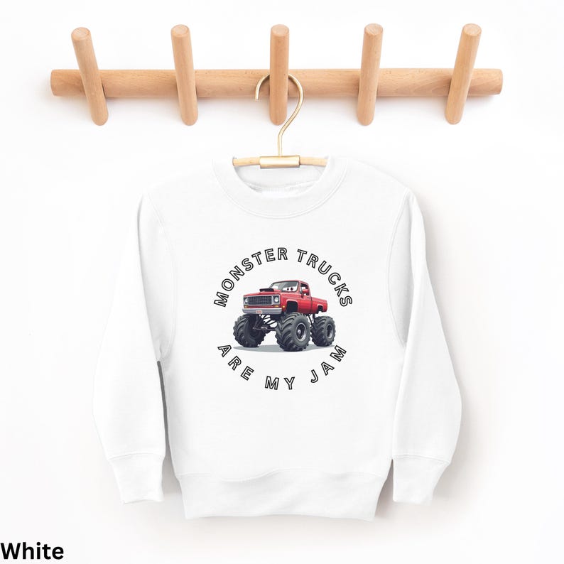 Puede incluir: Sudadera blanca con un gr&aacute;fico de cami&oacute;n monstruo rojo. El gr&aacute;fico presenta un cami&oacute;n rojo con neum&aacute;ticos negros grandes y el texto "Monster Trucks Are My Jam" en un dise&ntilde;o circular. La sudadera est&aacute; colgada en una percha de madera.