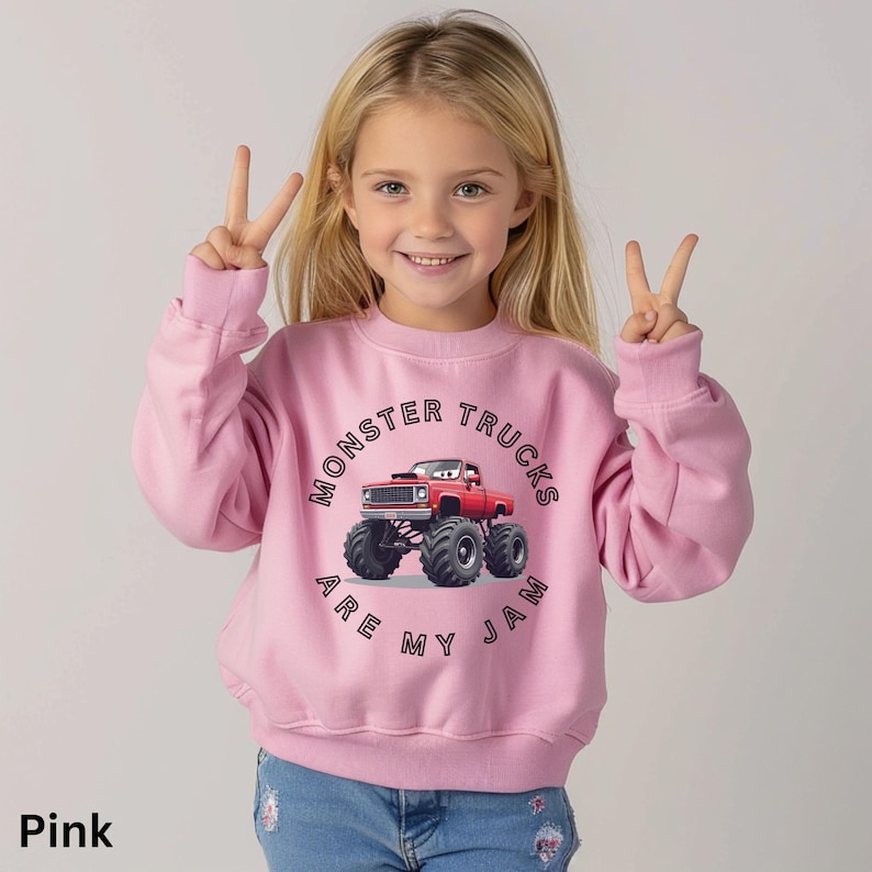 Puede incluir: Una sudadera rosa con un gr&aacute;fico de cami&oacute;n monstruo rojo y el texto "MONSTER TRUCKS ARE MY JAM". La sudadera la lleva una ni&ntilde;a. La ni&ntilde;a sonr&iacute;e y hace un signo de paz con los dedos.