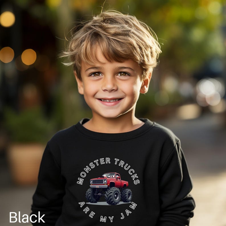 Puede incluir: Sudadera negra con un dise&ntilde;o de cami&oacute;n monstruo rojo y el texto "MONSTER TRUCKS ARE MY JAM". La sudadera la lleva un ni&ntilde;o peque&ntilde;o. El fondo est&aacute; borroso con tonos verdes y amarillos.