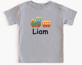 Könnte beinhalten: Ein graues T-Shirt mit einem bunten Zug-Motiv und dem Namen "Liam" in Schwarz. Der Zug hat rote, grüne, gelbe und blaue Details. Das Shirt besteht aus Athletic Heather Material.