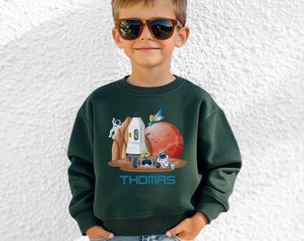 May include: A dark green sweatshirt featuring a colorful space-themed graphic. The design includes a rocket, astronauts, a red planet, and the name "THOMAS" in blue. The sweatshirt is worn by a child wearing sunglasses.