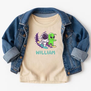 May include: A beige t-shirt featuring a graphic of an astronaut and a green alien surfing. The word "WILLIAM" is printed in teal below the image. The t-shirt is paired with a blue denim jacket.