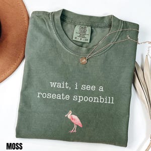 May include: A moss green crew neck t-shirt featuring the white text "wait, i see a roseate spoonbill" and a small illustration of a pink roseate spoonbill bird. The shirt has a Comfort Colors brand label inside the collar and a short gold chain.