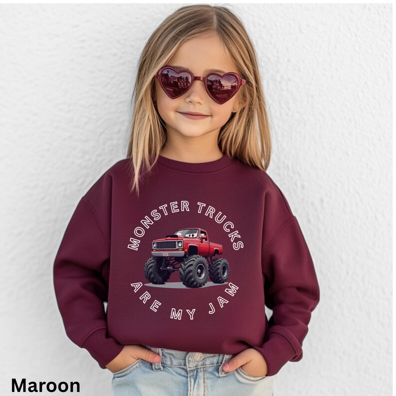Puede incluir: Sudadera granate con un gr&aacute;fico de cami&oacute;n monstruo rojo y el texto "MONSTER TRUCKS ARE MY JAM". La sudadera es usada por un ni&ntilde;o que lleva gafas de sol en forma de coraz&oacute;n.