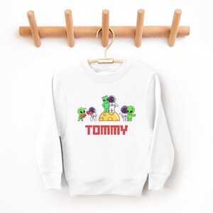 May include: White sweatshirt with a colorful graphic of aliens and astronauts on a yellow moon. The word "TOMMY" is printed in red block letters below the graphic. The sweatshirt is hanging on a wooden hanger.