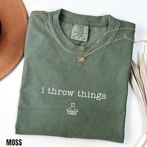 Puede incluir: Una camiseta verde musgo con la frase "i throw things" impresa en blanco. La camiseta tiene cuello redondo y mangas cortas. Un collar dorado con un colgante está drapeado sobre la camiseta. La camiseta está doblada y expuesta sobre una superficie blanca.