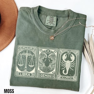 Könnte beinhalten: Ein moosgrünes T-Shirt mit einem weißen Grafikdruck von drei Tarot-Karten, die die Sternzeichen Waage, Zwillinge und Skorpion darstellen. Das T-Shirt hat einen Rundhalsausschnitt und ein Comfort Colors Markenetikett am inneren Kragen.