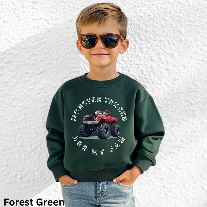 Puede incluir: Sudadera verde bosque con el texto "MONSTER TRUCKS ARE MY JAM" en un dise&ntilde;o circular blanco. El gr&aacute;fico presenta un cami&oacute;n monstruo rojo. La sudadera la lleva un ni&ntilde;o con gafas de sol.