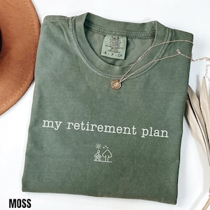 May include: A moss green short-sleeved t-shirt featuring the white text "my retirement plan" printed on the chest. Below the text is a small white line-art graphic of a person walking towards a tree under a sun and birds. The shirt has a crew neckline.