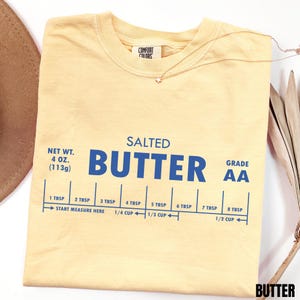 May include: A pale yellow short-sleeve t-shirt featuring blue text that reads SALTED BUTTER, NET WT. 4 OZ. (113g), GRADE AA, and a measurement guide ranging from 1 TBSP to 8 TBSP and 1/4 CUP to 1/2 CUP with the text START MEASURE HERE.