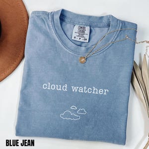 Könnte beinhalten: Ein blaues Jeans-T-Shirt mit den weißen Aufdruck "cloud watcher". Unter dem Text befinden sich drei weiße Wolkenillustrationen. Eine goldene Halskette mit Anhänger liegt über dem Shirt.